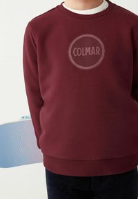 Lapsi, jolla on päällään marooninvärinen huppari, jossa on "COLMAR"-logo, pitää siniväristä skeittilautaa selkänsä takana.