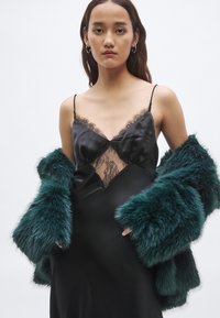 Μαύρο σατέν φόρεμα με λεπτομέρεια δαντέλας, συνοδευόμενο από ένα τιρκουάζ ριχτάρι από ψεύτικη γούνα. Το φόρεμα έχει λεπτές τιράντες και λαιμόκοψη σε σχήμα V.