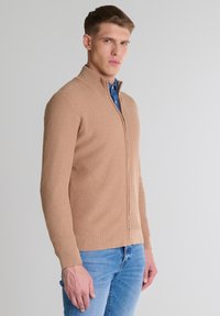 Beżowy sweter z prążkowanej tkaniny z zamkiem błyskawicznym, wysoki kołnierz, dopasowany krój i długie rękawy, w zestawie z niebieską koszulą na guziki pod spodem.