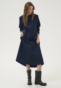 Rochie cămașă lungă, de culoare bleumarin, cu mâneci lungi, închidere cu nasturi, buzunare laterale și tiv asimetric, purtată cu cizme negre până la genunchi.