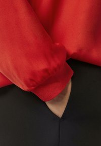 Rotes Satin-Oberteil mit langen Ärmeln und elastischen Bündchen, kombiniert mit einem schwarzen, figurbetonten Unterteil, das eine Seiten tasche und eine strukturierte stoffoberfläche aufweist.