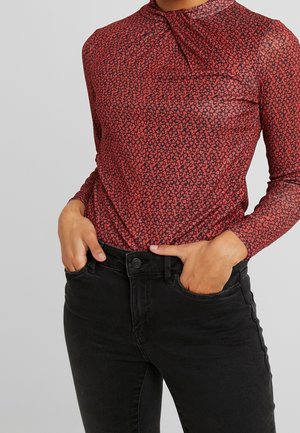 Czerwona bluzka z długim rękawem z drobnym kwiatowym wzorem, wpuszczona w czarne, wysokowalne dżinsy, ręce w przednich kieszeniach.