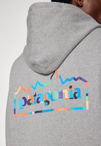 Pilkos spalvos džemperis su spalvingu spausdintu „patagonia“ ir kalnų dizainu ant nugaros, su tekstūruotu audiniu ir raišteliu kapuce.