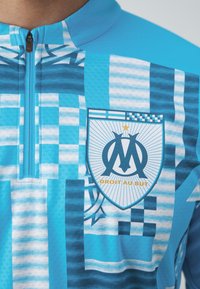 Maglia sportiva blu con colletto a mezza zip, motivo grafico e emblema del club con il testo "DROIT AU BUT" in colori a contrasto.