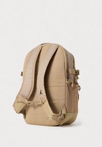 Mochila castanha com costas almofadadas, alças ajustáveis, bolso lateral em rede e um pompom decorativo. Apresenta um pequeno logo na parte da frente.