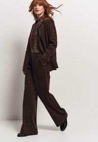 Completo in velluto a coste marrone con pantaloni a gamba larga e giacca sartoriale. Sotto, un top aderente con stampa leopardata aggiunge contrasto. Scarpe nere completano l'outfit.