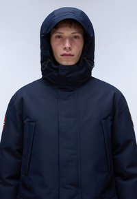 Giacca isolata di colore blu navy con collo alto e cappuccio regolabile, dotata di tasche laterali e tessuto testurizzato. Design semplice con accenti minimal.