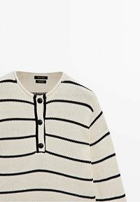 Cremefarbener Strickpullover mit schwarzen horizontalen Streifen und einer teilweisen Knopfleiste mit drei schwarzen Knöpfen auf der Brust.