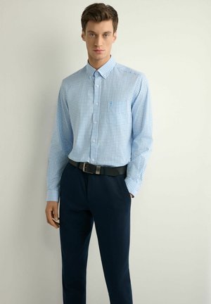 Mężczyzna w jasnoniebieskiej, kraciastej koszuli zapinanej na guziki, włożonej do ciemnogranatowych spodni z czarnym paskiem, stoi na tle jasnej, jednolitej ściany.