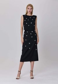 Robe noire sans manches avec une conception de haut court. Présente une broderie florale blanche et une jupe droite arrivant au genou.