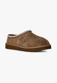 Zapatilla de ante marrón con un exterior texturizado, un ribete trenzado y una suela plana de goma. Presenta la marca UGG en el lateral.
