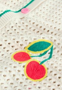 Tissu en maille blanche avec broderie de cerises rouges et de feuilles vertes soulignées de jaune, et un bouton rose sur le bord supérieur.