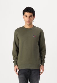 Maglione verde oliva in morbido materiale, con scollo rotondo e polsini a coste. Piccolo patch con logo sul lato sinistro del petto.