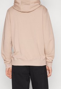 Sudadera beige de tejido suave, de corte holgado y con una gran capucha. Presenta puños y dobladillo de canalé, combinada con pantalones negros.