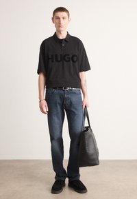 Juoda polo marškiniai su dideliu "HUGO" logotipu, mėlynos džinsinės kelnės, juodi loafersai ir juoda odinė rankinė, stovintys ant neutralios fono.