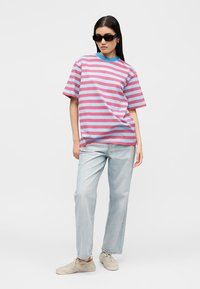 Γυναίκα στέκεται φορώντας υπερμεγέθη ροζ και μπλε ριγέ μπλουζάκι, ανοιχτό μπλε τζιν, μπεζ αθλητικά παπούτσια και μεγάλα μαύρα γυαλιά ηλίου σε λευκό φόντο.