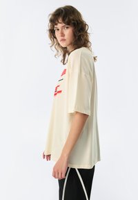 Crème-kleurige oversized t-shirt met korte mouwen, voorzien van rode grafische tekst en lijnontwerpen. Zachte textuur en een relaxte pasvorm.