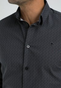 Homme portant une chemise à boutons sombre avec un petit motif géométrique répétitif et un col montant.
