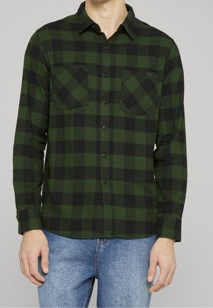 Homme portant une chemise à carreaux verte et noire avec deux poches poitrine et un jean bleu clair, debout devant un fond gris uni.