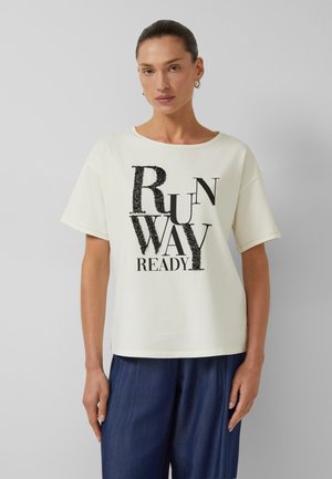 Kobieta z zaczesanymi do tyłu włosami nosi białą koszulkę z czarnym napisem „RUNWAY READY” oraz luźne ciemnoniebieskie spodnie na jednolitym tle.