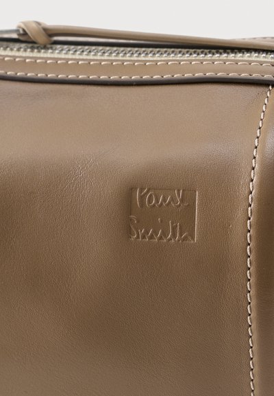Κοντινό πλάνο μπεζ δερμάτινης τσάντας με εμφανείς ραφές, ανάγλυφο λογότυπο Paul Smith και μερικώς ορατό μεταλλικό φερμουάρ στην πάνω άκρη.
