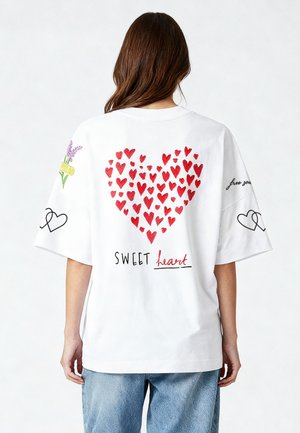 Valkoinen ylisuuressa t-paidassa on iso punainen sydäntaipe, teksti "SWEET heart" ja kukkakuosiset hihat. Pehmeä puuvillakangas.