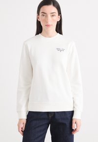 En kvinne med langt mørkt hår har på seg en kremfarget genser og mørke jeans, stående mot en enkel hvit bakgrunn.