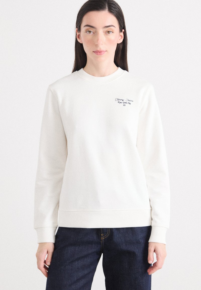 En kvinne med langt mørkt hår har på seg en kremfarget genser og mørke jeans, stående mot en enkel hvit bakgrunn.
