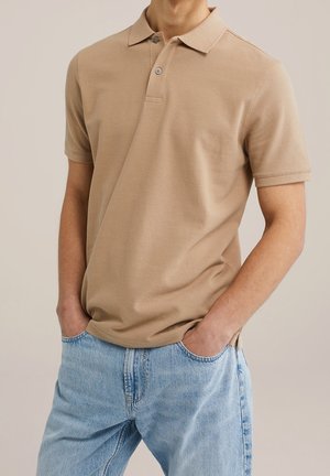 Beige kurzärmliges Polo-Shirt getragen zu hellblauen Jeans, Hände lässig in den vorderen Taschen, neutraler Hintergrund.