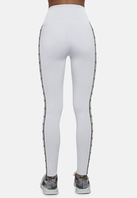 Białe legginsy z wysokim stanem, z czarnymi paskami po bokach i gładką powierzchnią. W zestawieniu z szarymi butami sportowymi.