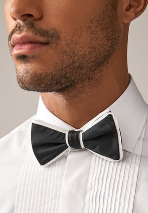 Mees hoolitsetud habemega, seljas valge särk ja must kikilips valgete äärtega, taustaks neutraalne varjund.