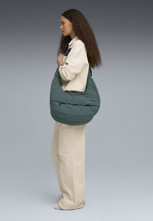 Gev Quilted donkerblauwe hobo tas met een ronde vorm, voorzien van een ritsvak aan de voorkant en een schouderband. Model draagt een lichte top en brede beige broek.