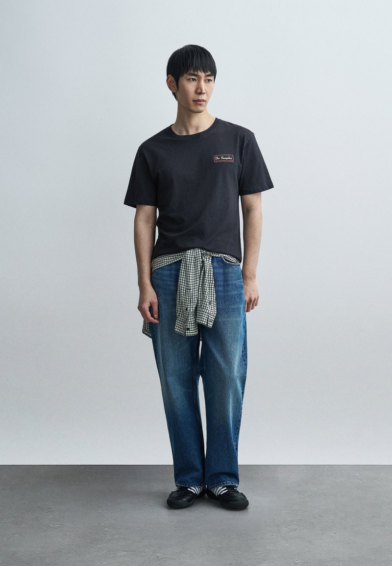 Black short-sleeve T-shirt with embroidered logo, paired with loose blue jeans and a tied black-and-white checkered shirt around the waist.