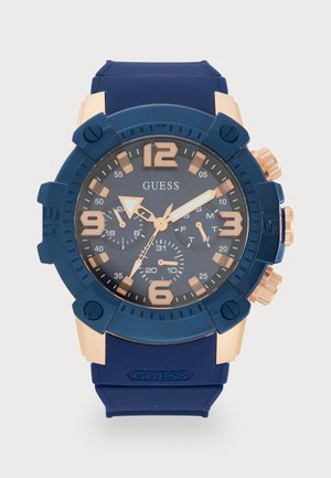 Reloj con correa de silicona azul, esfera redonda de color azul oscuro, acentos en tono dorado y marcadores numéricos grandes; presenta subesferas de cronógrafo y pulsadores.