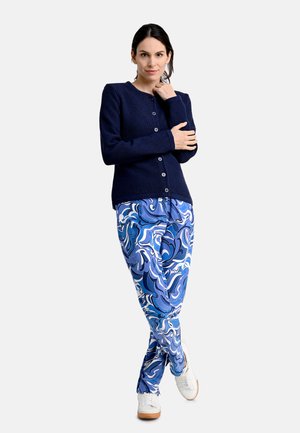 Marineblauer Strickcardigan mit Rundhalsausschnitt und Knopfverschluss, kombiniert mit blauen gemusterten Hosen mit Wirbeln in Blau und Weiß. Weiße Sneaker.
