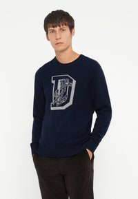 Pull marin en tricot de couleur marine, présentant une grande lettre "D" grise avec un motif d'ancre. Encolure et poignets côtelés, coupe décontractée.
