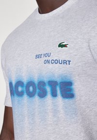 Grå bomulls-t-shirt med en grön krokodil-logga, blå text som säger "SE DIG PÅ PLANEN," och en gradient blå accent längst ner.