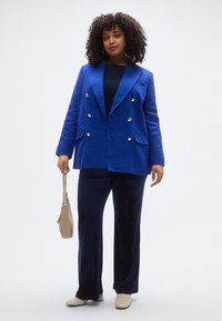 Μπλε βελούδινο σακάκι διπλής όψης με χρυσά κουμπιά, σε συνδυασμό με σκούρο μπλε παντελόνι φαρδιάς γραμμής. Το μοντέλο κρατά μια καφέ τσάντα και φορά ανοιχτόχρωμα παπούτσια.