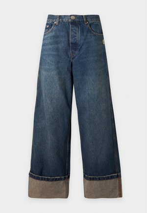 Jeune femme debout portant un t-shirt blanc sans manches Marc Jacobs et un jean bleu large avec revers, ainsi que des chaussures noires, sur un fond uni.