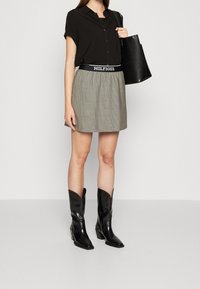 Schwarzes kurzärmeliges Hemd mit Knöpfen, kombiniert mit einem karierten Rock mit einem gebrandeten Bund. Schwarze Lederstiefeletten und eine Tragetasche runden den Look ab.