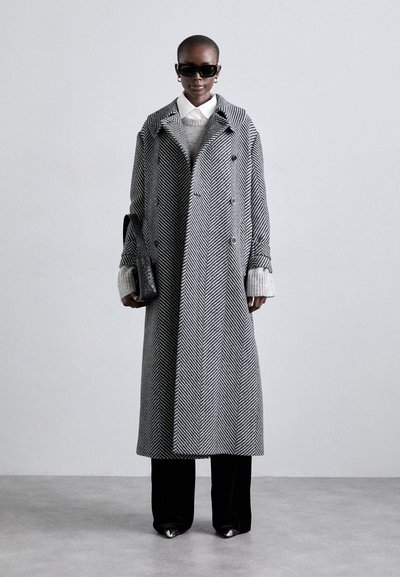 Doppelt belegter Fischgrätenmantel in Schwarz und Weiß, kombiniert mit einem grauen Pullover und schwarzen Hosen, accessoriert mit einer schwarzen Tasche und Sonnenbrille.