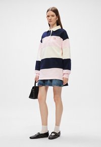 Μακρυμάνικο πόλο με ρίγες σε ναυτικό, ροζ και κρέμα χρώμα; χαλαρή εφαρμογή. Συνδυασμένο με τζιν φούστα και μαύρη τσάντα, με μαύρα παπούτσια και ανοιχτόχρωμες κάλτσες.