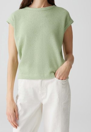 Femme portant un haut en maille vert clair à manches courtes et un pantalon blanc taille haute, une main dans la poche, debout devant un fond uni.