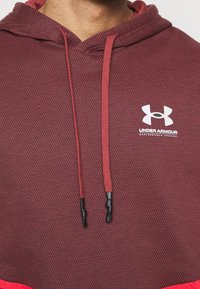 Roter und dunkelroter Hoodie aus einem strukturierten Stoff. Verfügt über eine Kapuze, Kordelzüge mit schwarzen Spitzen und ein weißes Under Armour-Logo auf der Brust.