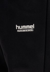 Sort sportstøjsstof med hvidt "hummel"-logo og teksten "SPORTSWEAR", med en glat tekstur og struktureret design.