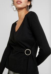 Černý pletený svetr s hlubokým V-výstřihem a šroubovaným designem, který má po straně zlatý prsten jako stylový detail.