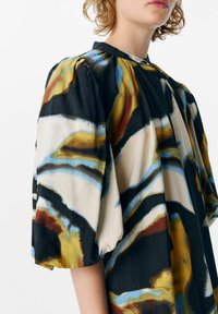 Blouse à imprimé floral avec des manches bouffantes, un col montant, et un mélange de couleurs incluant le noir, le bleu, l'or et le rouge sur un fond clair.
