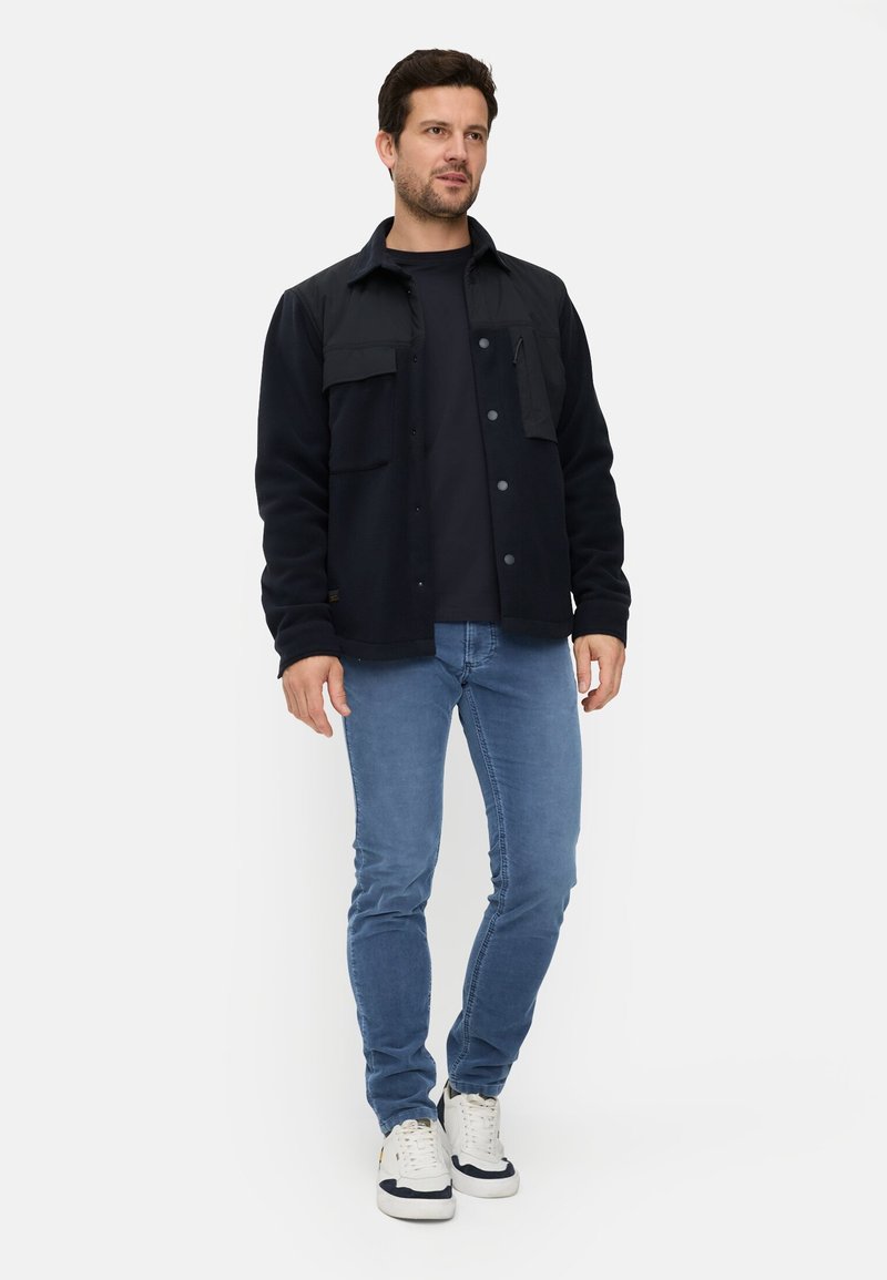 Schwarzes Fleece-Oberhemd mit Knopfleiste und Taschen, kombiniert mit einem schwarzen T-Shirt, hellblauen Jeans und weißen Turnschuhen.