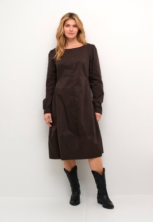 Bruine jurk met lange mouwen, rechte silhouet, gestructureerde stof, verzamelde mouwen en een lengte tot de knie. Draag met zwarte cowboylaarzen.