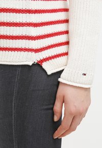 Gestrickter Pullover mit rot-weißen horizontalen Streifen; gerippte Bündchen; seitliche Schlitze; Logo auf dem Ärmel gestickt; strukturierter Stoff.
